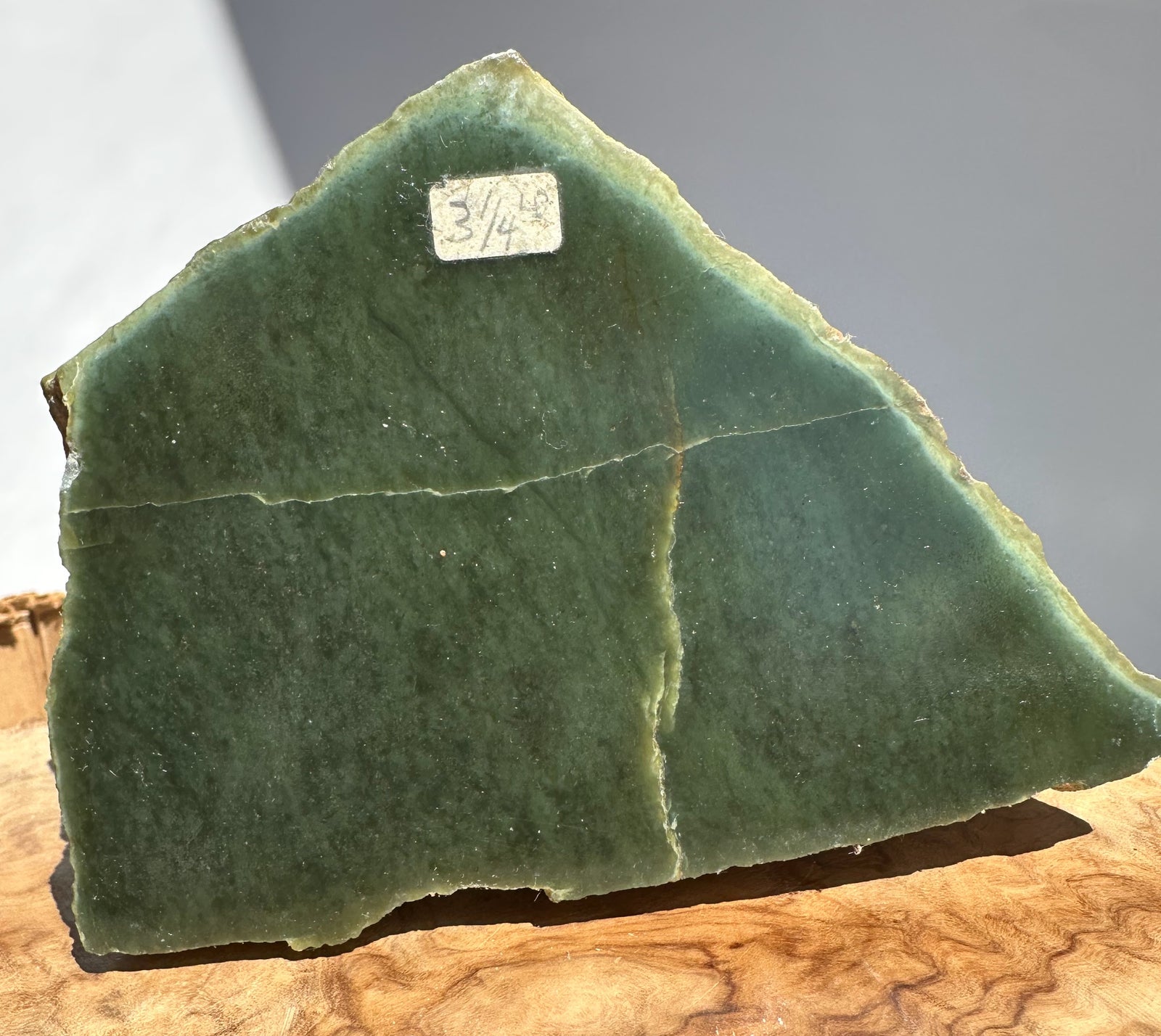 A raw green jade with natural fissures and rough edges, resting on a piece of wood against a neutral gray background. A small handwritten price tag is visible on the top edge.
