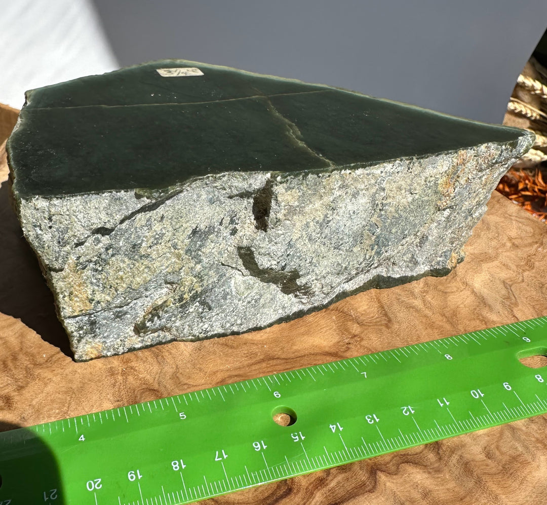 A raw dark green and gray stone slab with natural fissures, shown next to a green measuring ruler on a wood-grain surface.