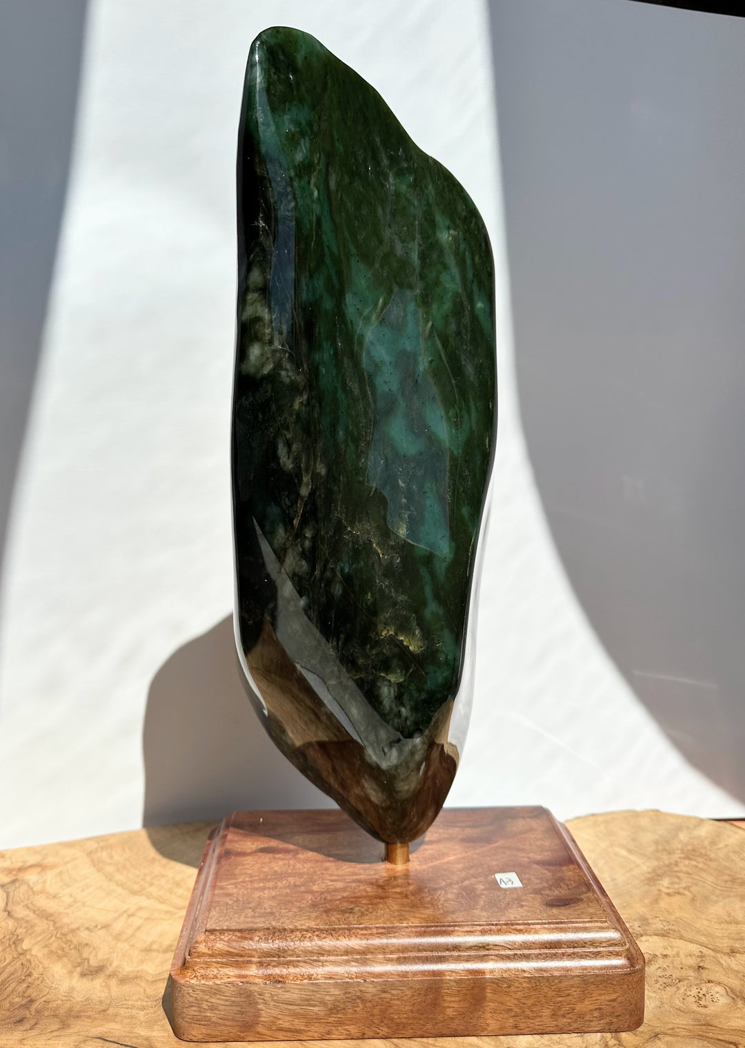 A polished dark green stone sculpture with natural marbling, mounted on a rectangular wooden base, photographed indoors against a white wall with strong sunlight and shadows.