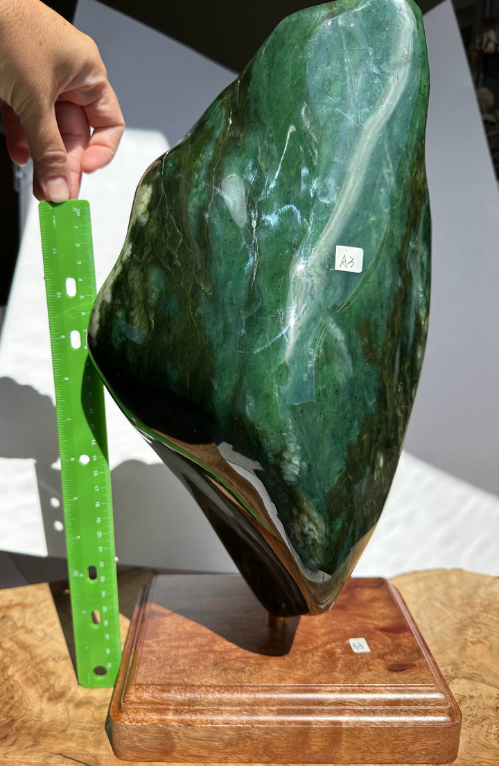 A polished green jade sculpture on a wooden display base, with a hand holding a green ruler beside it for scale. The stone has glossy highlights and natural marbling, photographed indoors against a light background.
