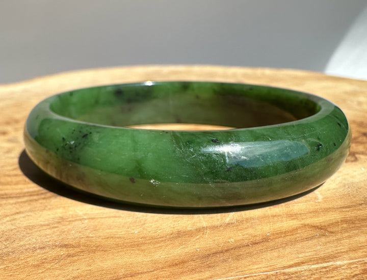 Front-angle shot of a green nephrite jade bangle with dark speckles on a wooden surface.