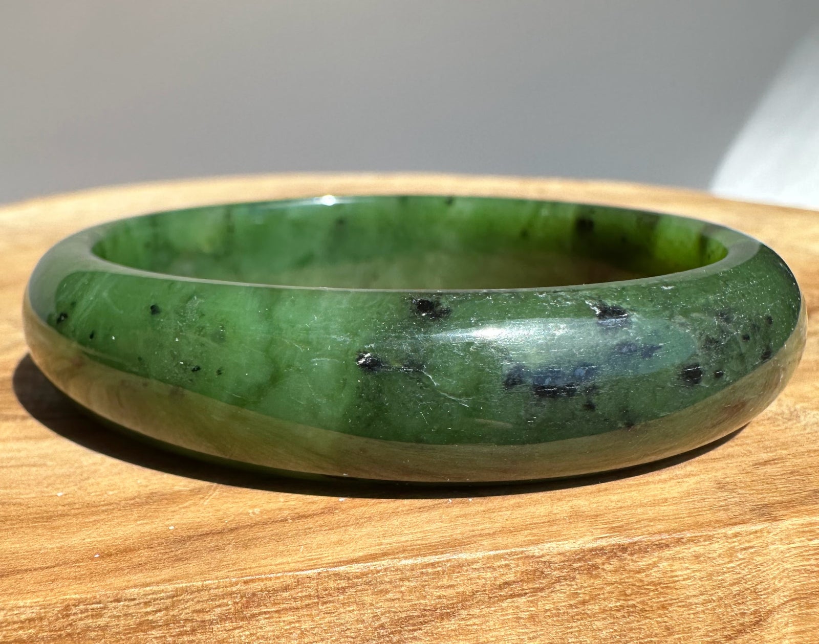 Green nephrite jade bangle with black inclusions shown on a wooden surface