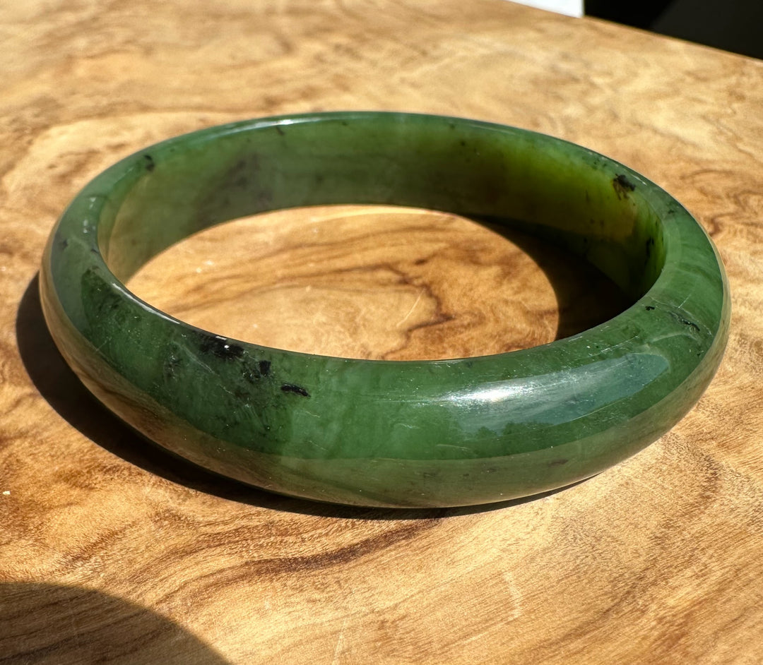 Close-up of a green nephrite jade bangle on a wood surface showing the inner edge and dark inclusions.