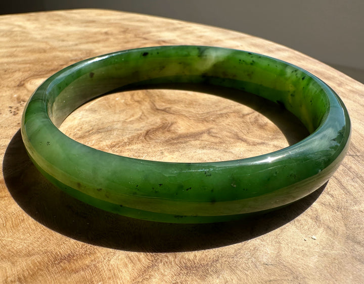 Top angle of jade bangle with visible dark mineral inclusions and translucent green hues on wood.