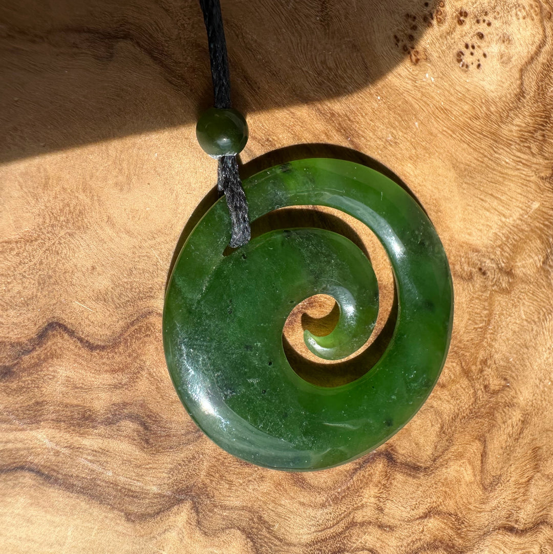 Green Canadian Jade spiral pendant with a small round bead on a black cord against a wooden background.
