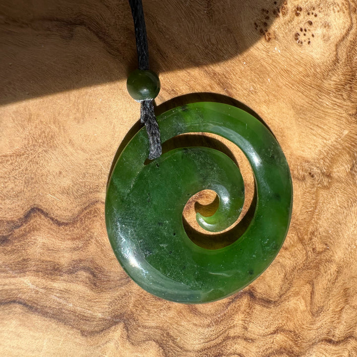 Green Canadian Jade spiral pendant with a small round bead on a black cord against a wooden background.