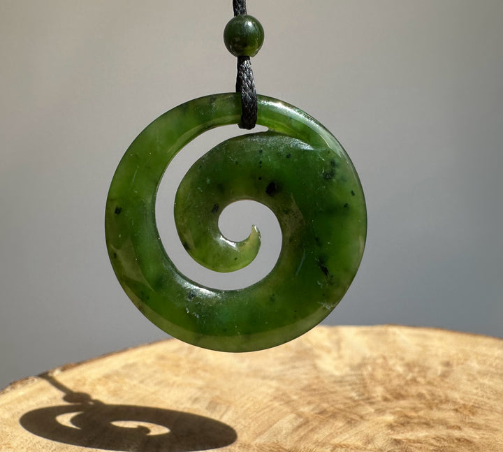 Green Canadian Jade pendant with a round bead above, suspended on a black cord against a neutral gray backdrop.
