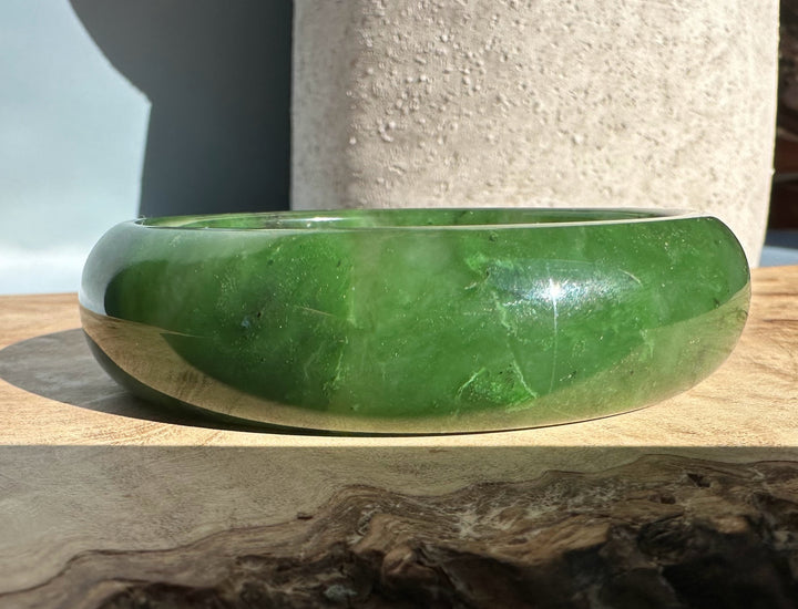 Canadian Jade Bangle 58 x 17.5mm in rich green with a smooth rounded edge resting on wood under bright light