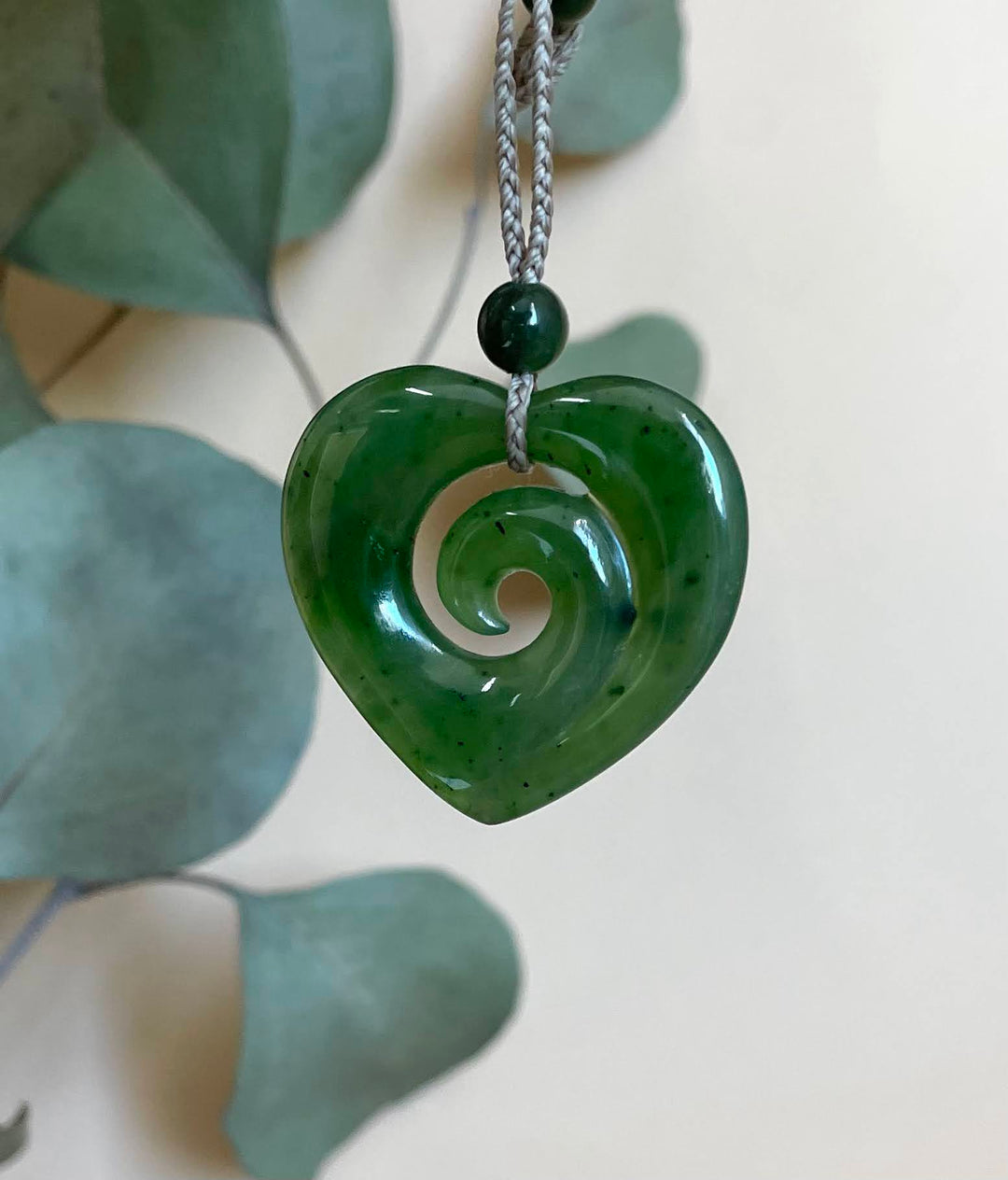 A green carved stone heart pendant with a spiral cutout, hanging from a gray braided cord with a dark green bead, set against a soft beige background with blurred green leaves.