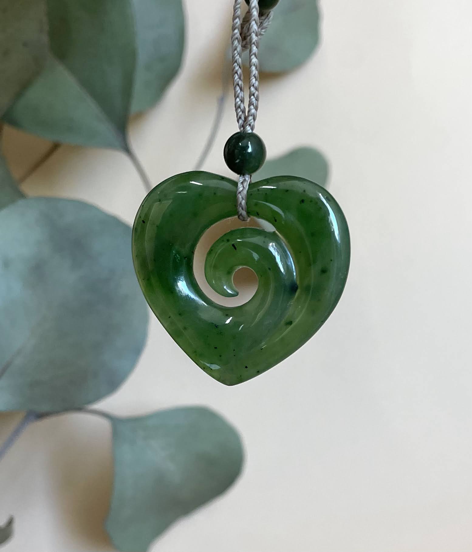 A green carved stone heart pendant with a spiral cutout, hanging from a gray braided cord with a dark green bead, set against a soft beige background with blurred green leaves.