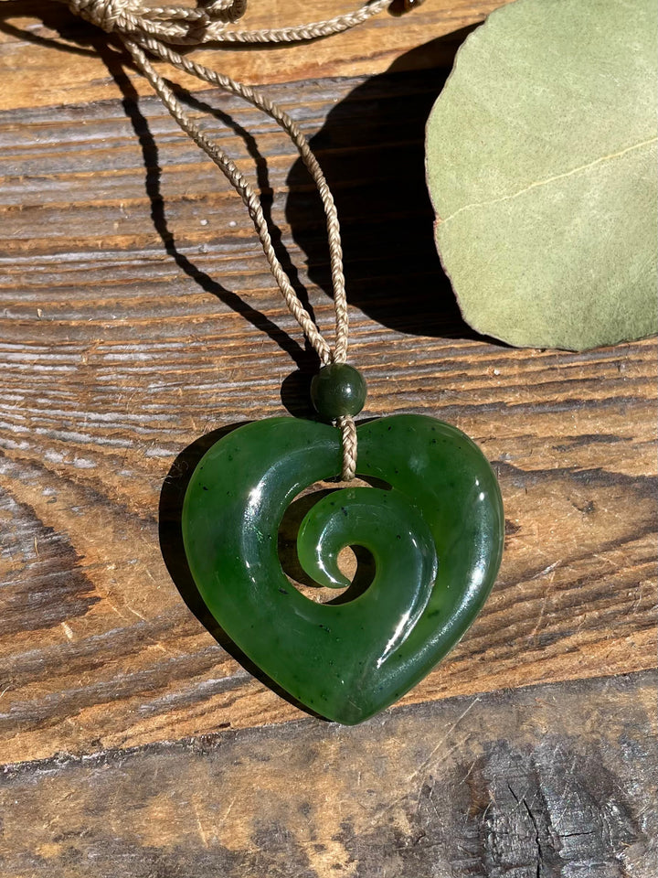 A dark green carved heart-shaped pendant necklace with a spiral motif, hanging on a natural fiber cord over a rustic wooden surface.