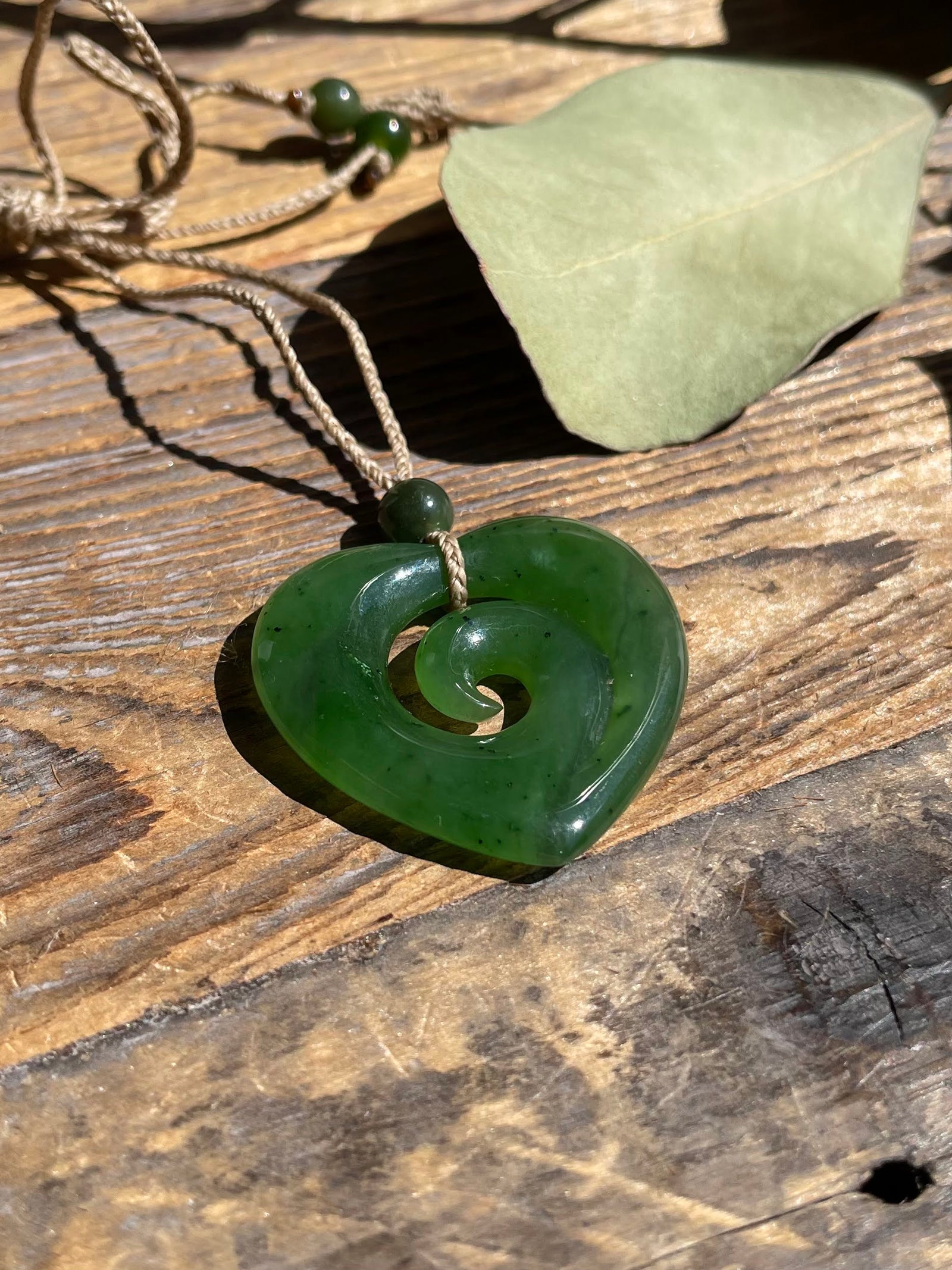A green carved stone pendant necklace with a spiral cutout, hanging from a tan cord on a rustic wooden surface with a dried leaf nearby.