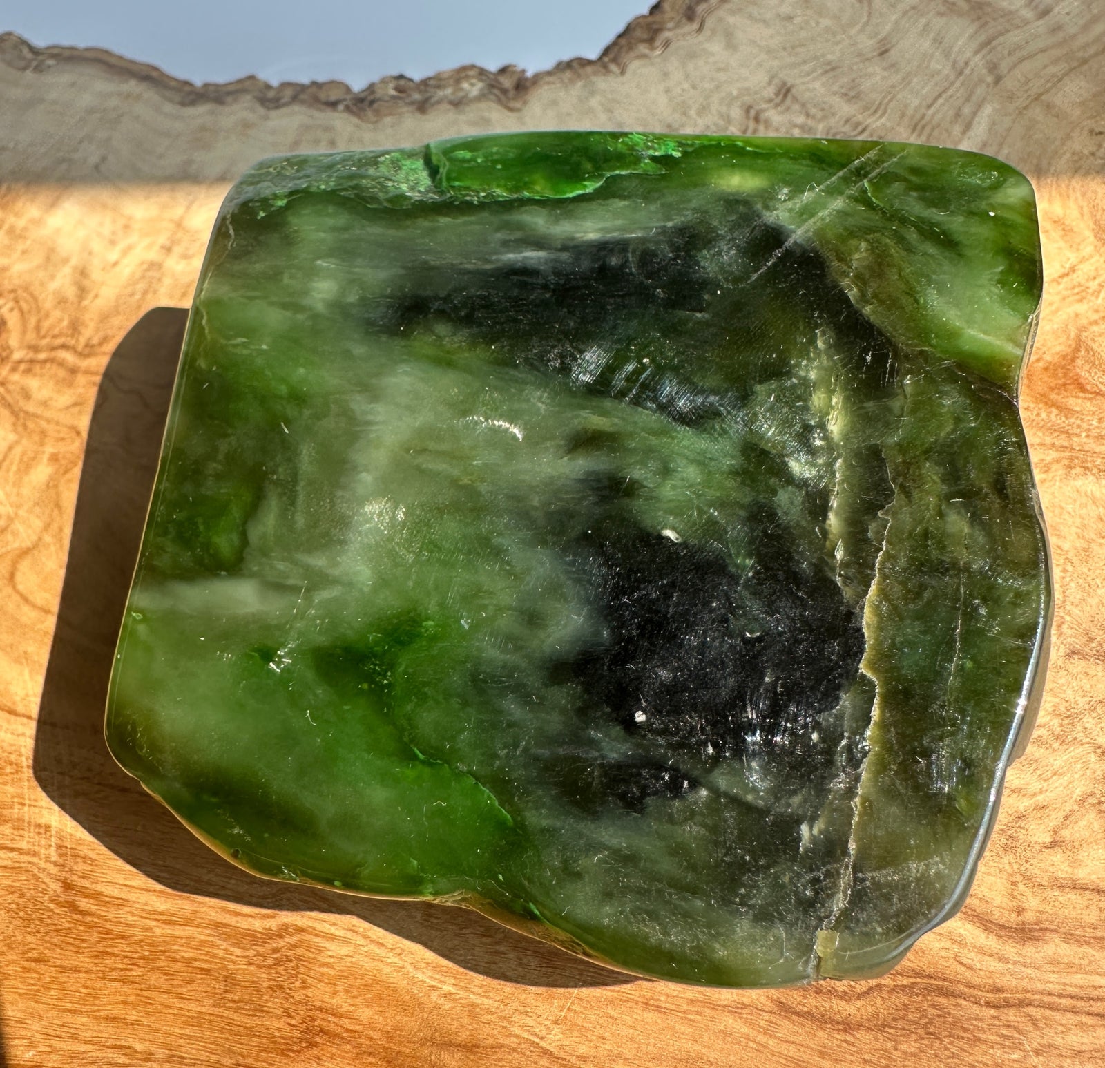 A polished green jade slab with natural marbling and dark inclusions, displayed on a curved wooden surface with sunlight casting a shadow.