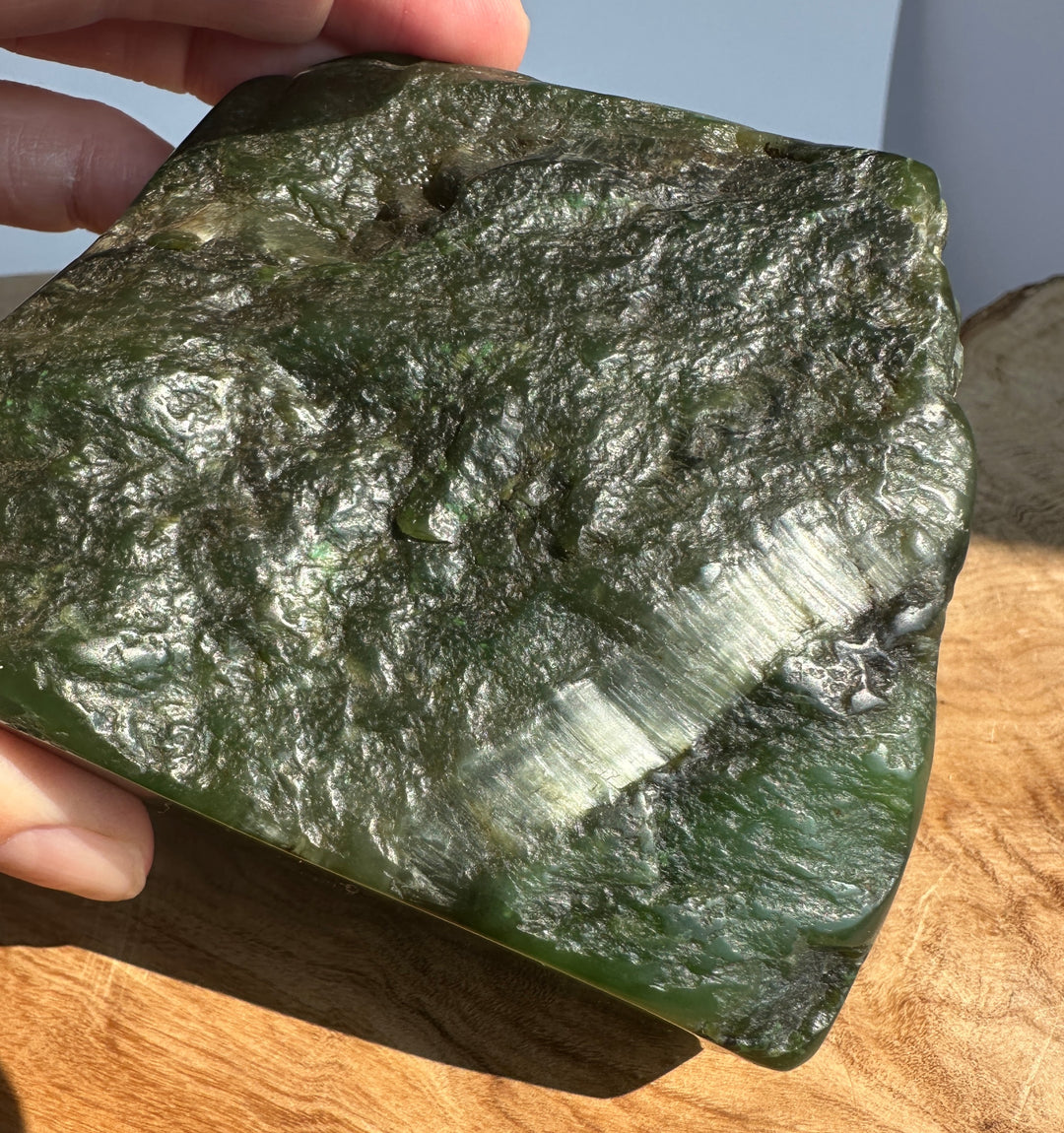 A hand holds a rough, polished green jade slab with natural marbled texture and a bright reflective streak, photographed over a wooden surface in natural light.