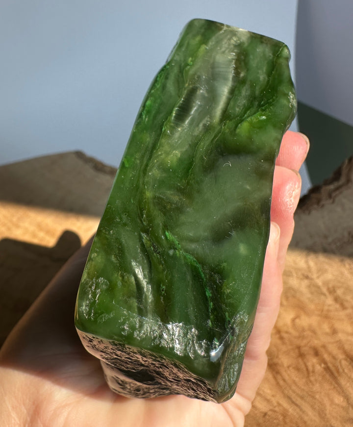 A hand holding a polished green jade with a rough dark base, photographed indoors against a soft blue-gray background.