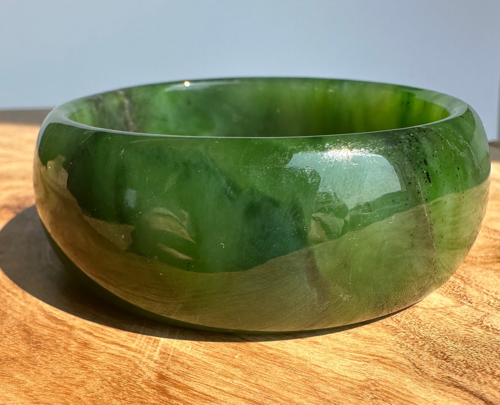 A polished green jade bangle bracelet displayed on a wooden surface with a light blue background.
