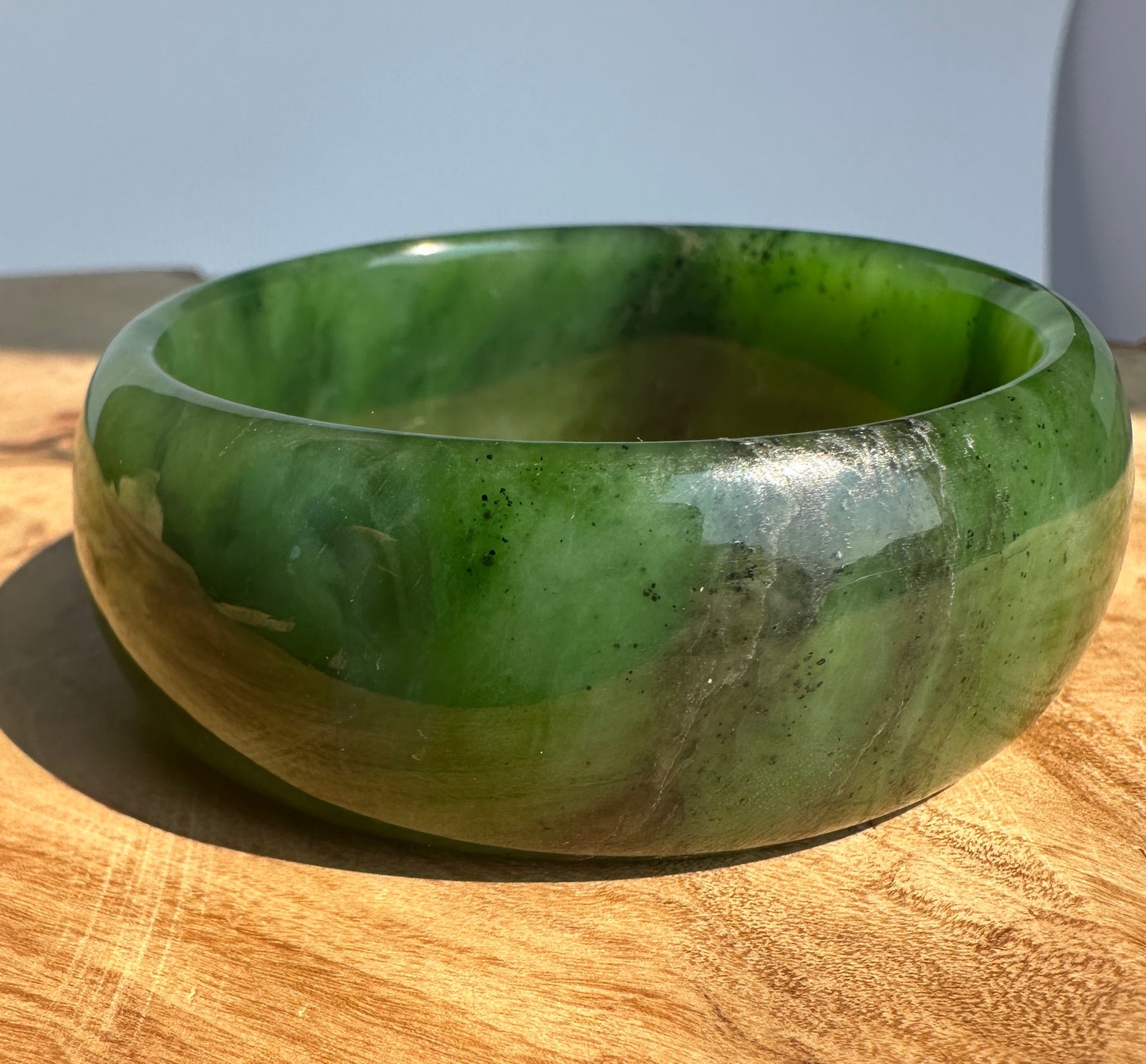 A polished green jade bangle bracelet resting on a wooden surface under natural sunlight.