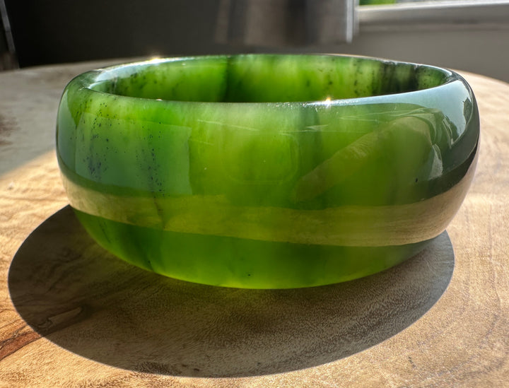 A thick, wide green stone bangle with natural marbled patterns, resting on a wooden surface in natural sunlight.