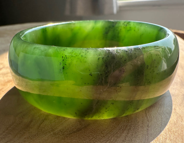 A round green jade bangle bracelet with natural inclusions and a polished finish, resting on a wooden surface.