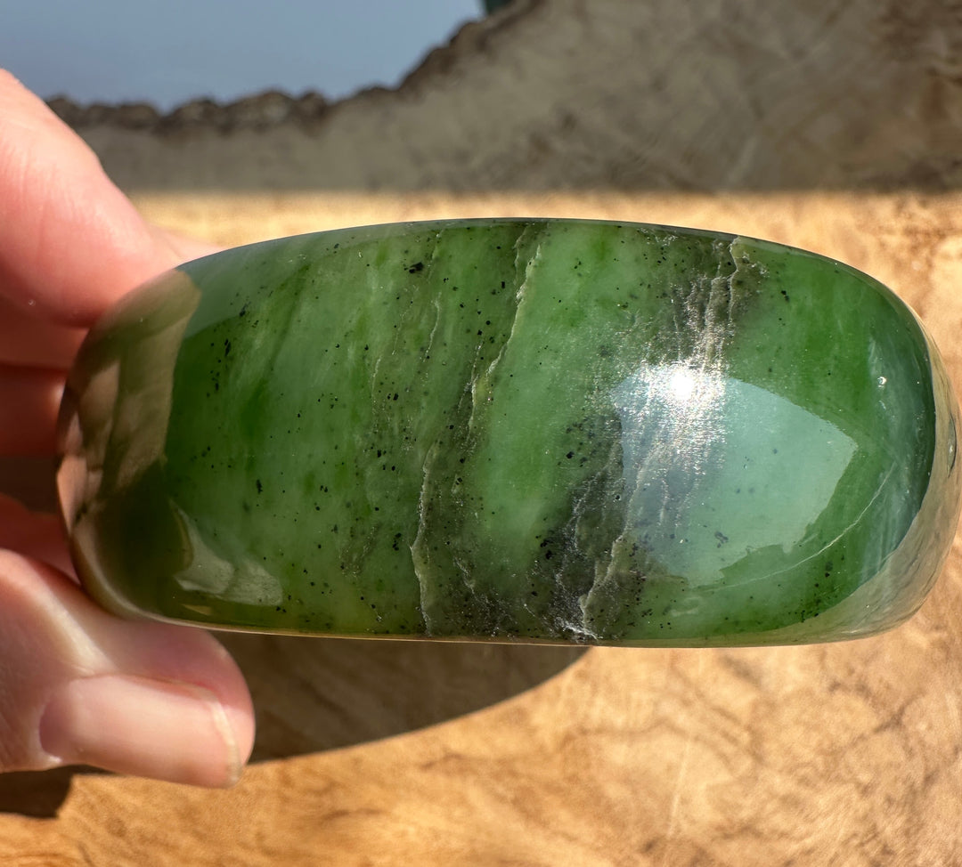 A hand holding a polished green jade bangle bracelet with natural inclusions and cloudy white veining, set against a rustic wooden background.