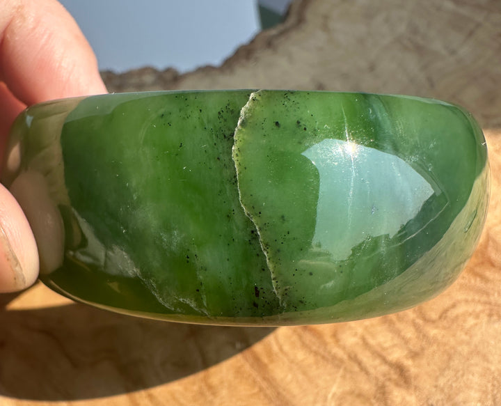 A hand holding a polished green jade bangle bracelet against a natural wood and outdoor background.
