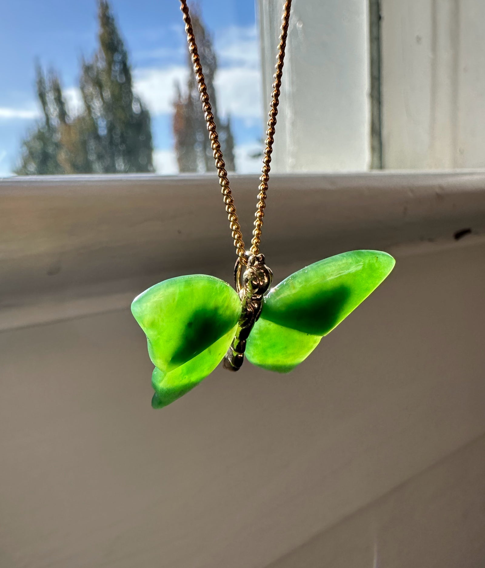 Green jade butterfly pendant on a gold chain hanging near a sunlit window