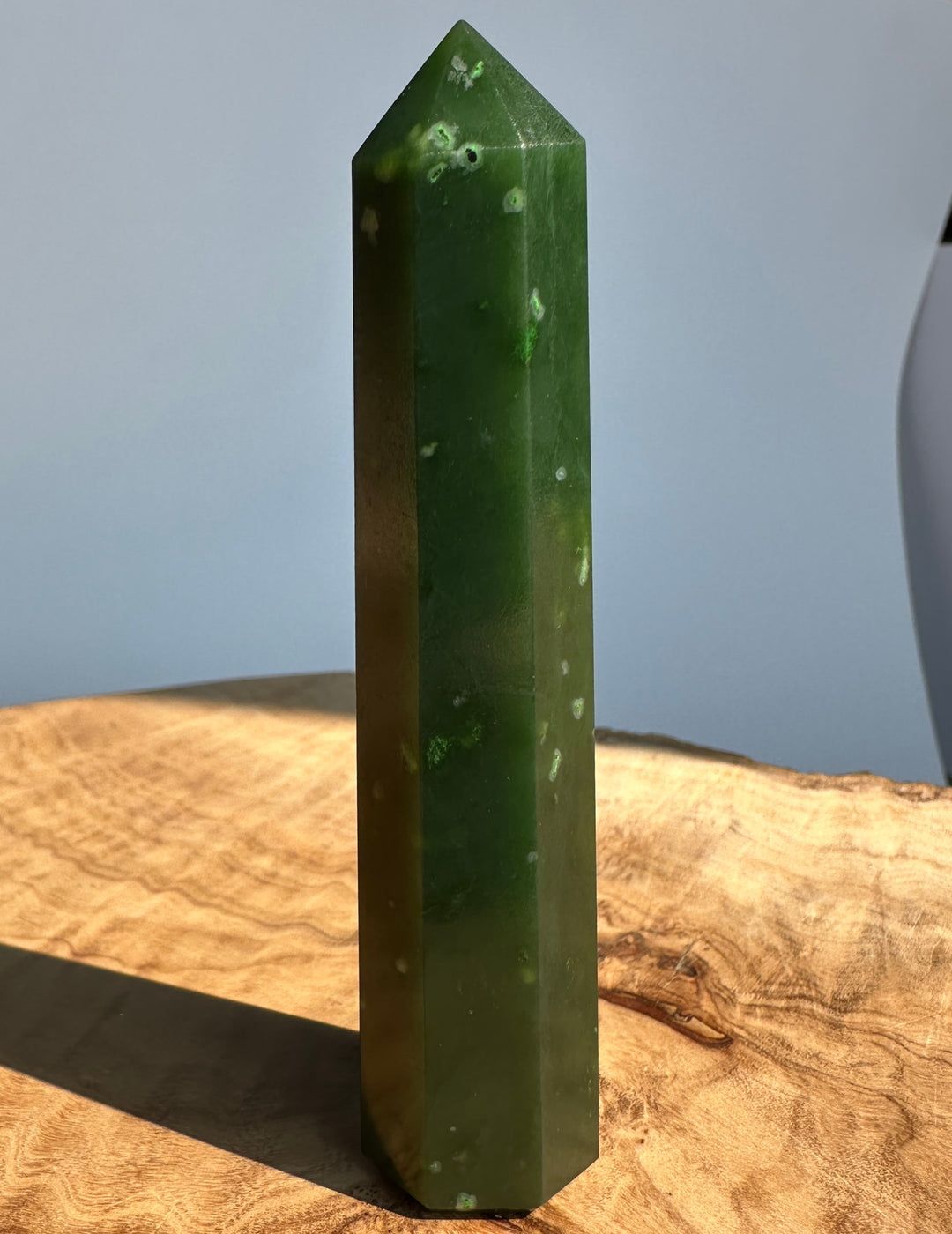 A polished dark green crystal tower with natural white inclusions, standing upright on a rustic wooden surface against a soft gray background.