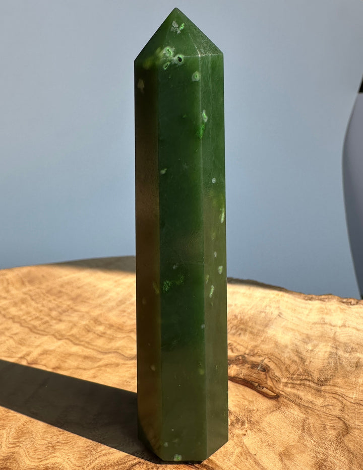 A polished dark green crystal tower with natural white inclusions, standing upright on a rustic wooden surface against a soft gray background.