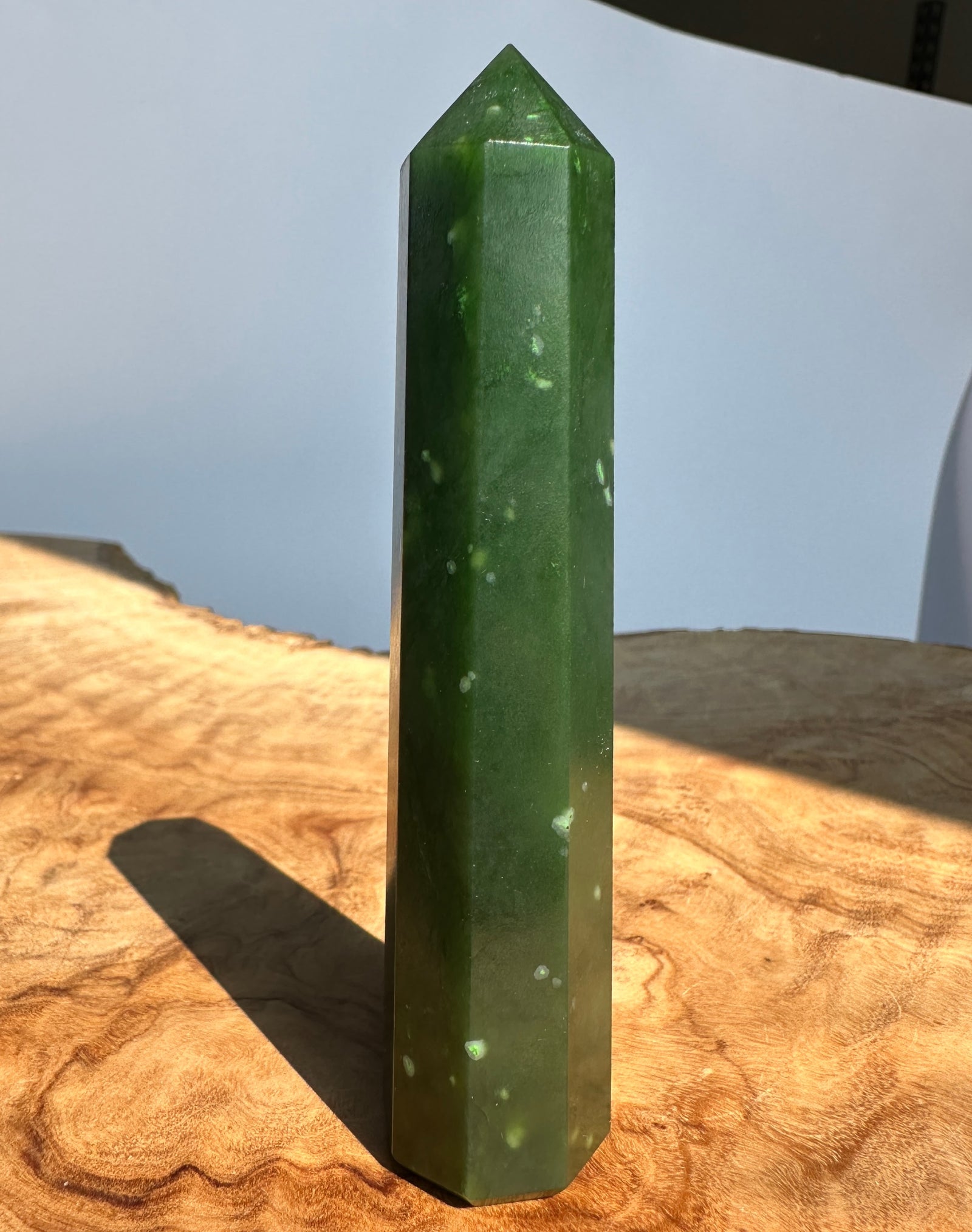A polished green jade crystal tower with a pointed top, standing upright on a natural wood slab with a soft-focus outdoor background.