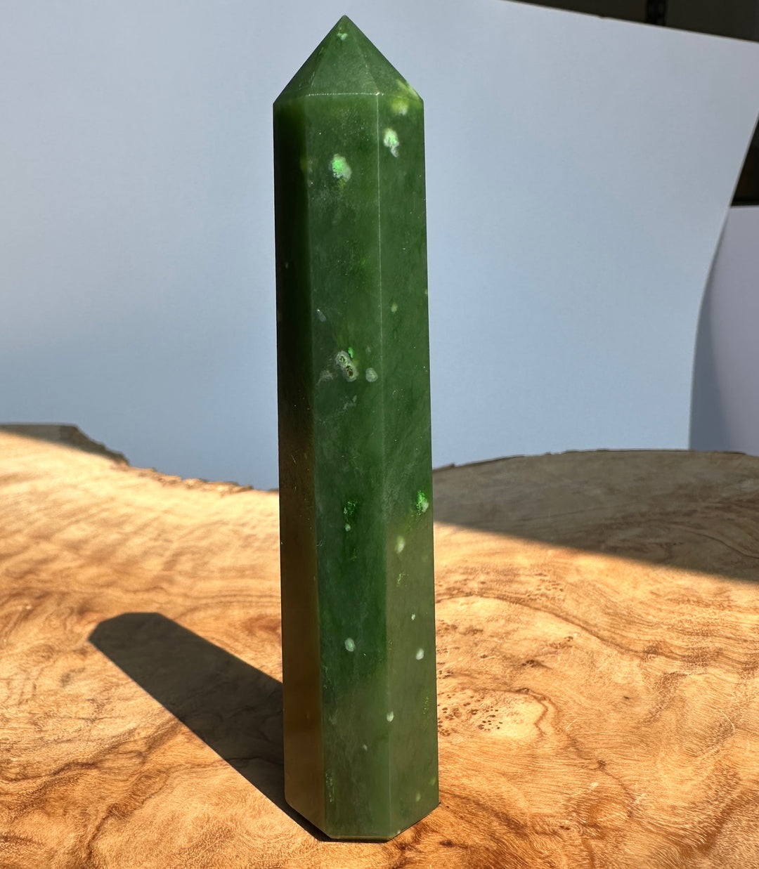 A polished green crystal obelisk with white inclusions, standing upright on a rustic wood slice surface with a soft-focus light blue background.