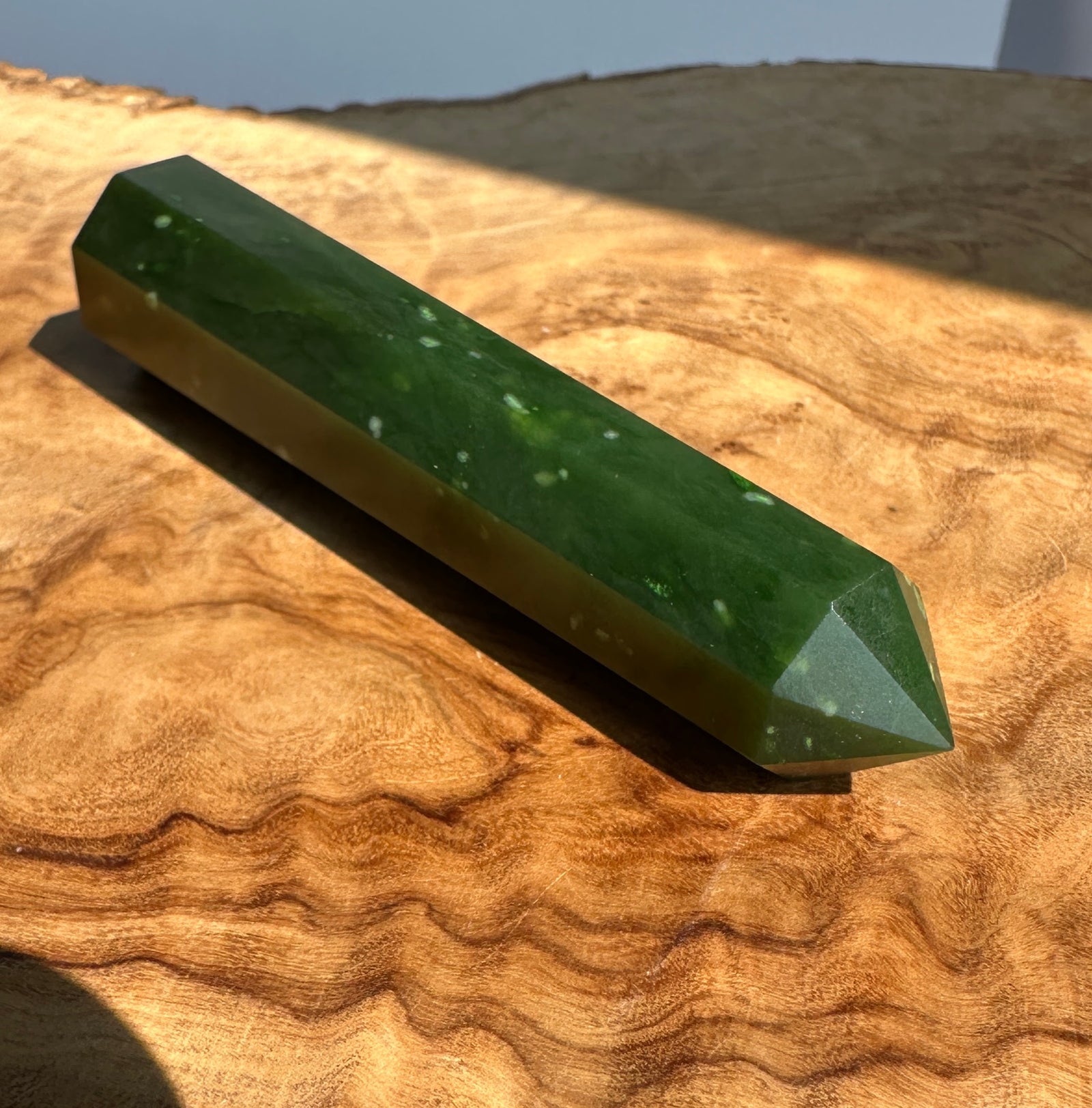 A polished green jadel point stake resting diagonally on a textured, wavy wood slice surface under bright natural sunlight.