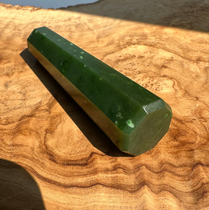 A green faceted crystal wand resting diagonally on a textured, wavy wood surface under bright natural sunlight.