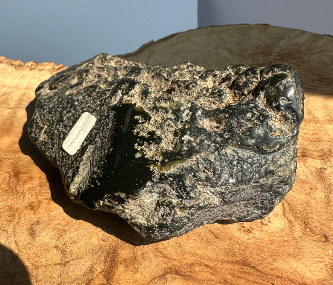A rough dark jade stone or mineral specimen with a small white label, resting on a curved piece of natural wood against a pale blue-gray background.