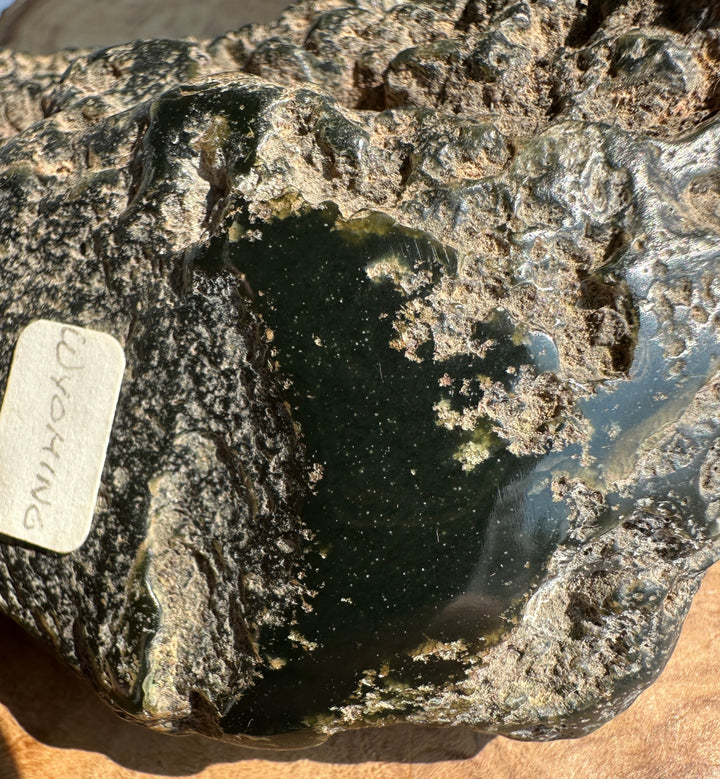 A polished dark green jade  with rough natural edges and a small white handwritten label attached, photographed close up on a tan surface.