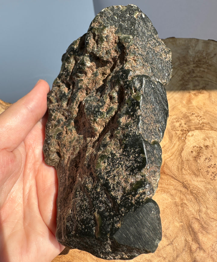 A hand holding a rough, dark green and brown jade stone or mineral specimen against a neutral background with a wood surface below.