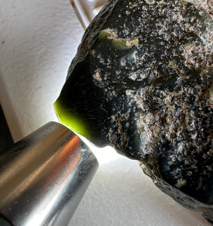 A close-up of a rough dark stone or crystal being examined with a bright light, showing a vivid yellow-green internal inlay or cavity. A metal light source is visible at the lower left against a white background.