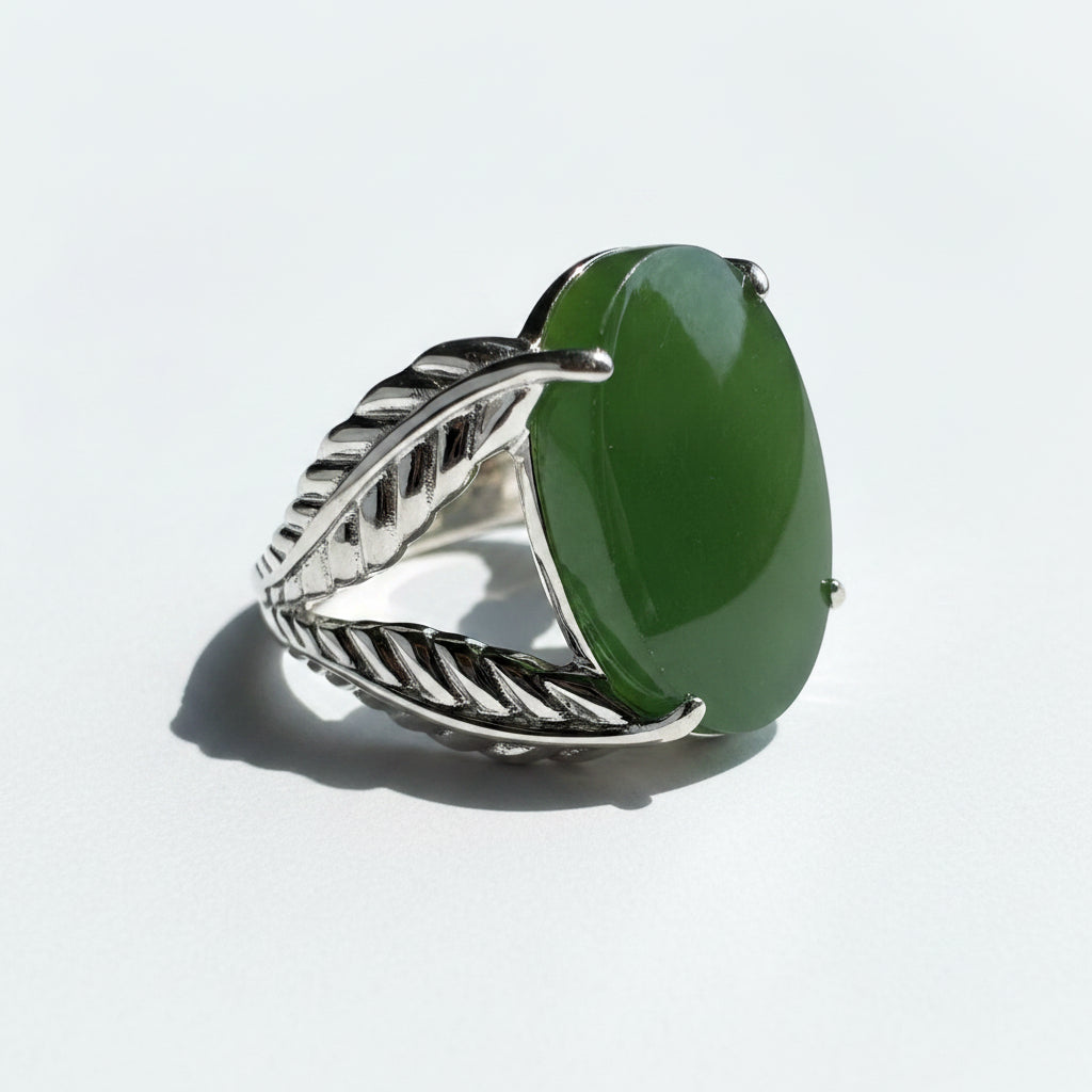 A silver-toned ring designed like a leaf, featuring a large, smooth green stone on the front, resting on a white surface.