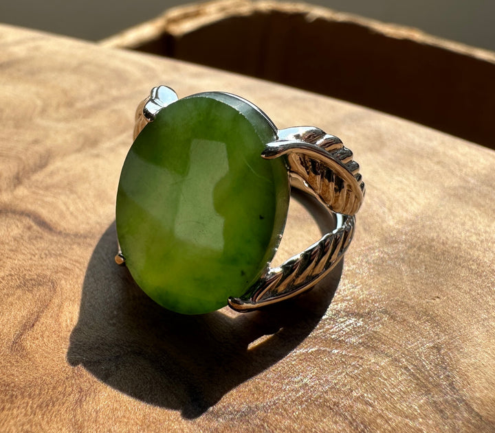 A silver-toned ring with a large oval green stone, photographed close-up on a wooden surface with warm natural light and a soft shadow.
