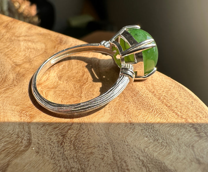 A silver ring with a round green gemstone in a cage-like setting, resting on a textured wooden surface with warm sunlight and shadows.
