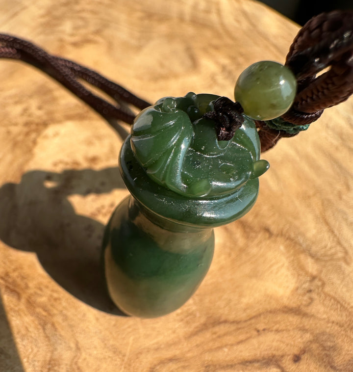 A green jade pendant with a carved bat top and a dark brown cord, resting on a wooden surface.