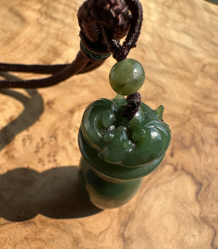 A carved green jade pendant with a dark brown cord and a round green bead, resting on a textured wooden surface.