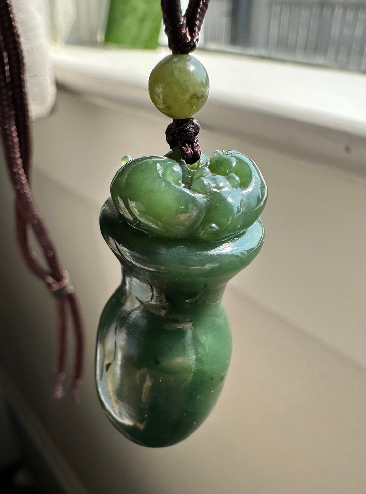 A green jade pendant c, hanging from a dark cord with a green bead, set against a blurred indoor background near a window.