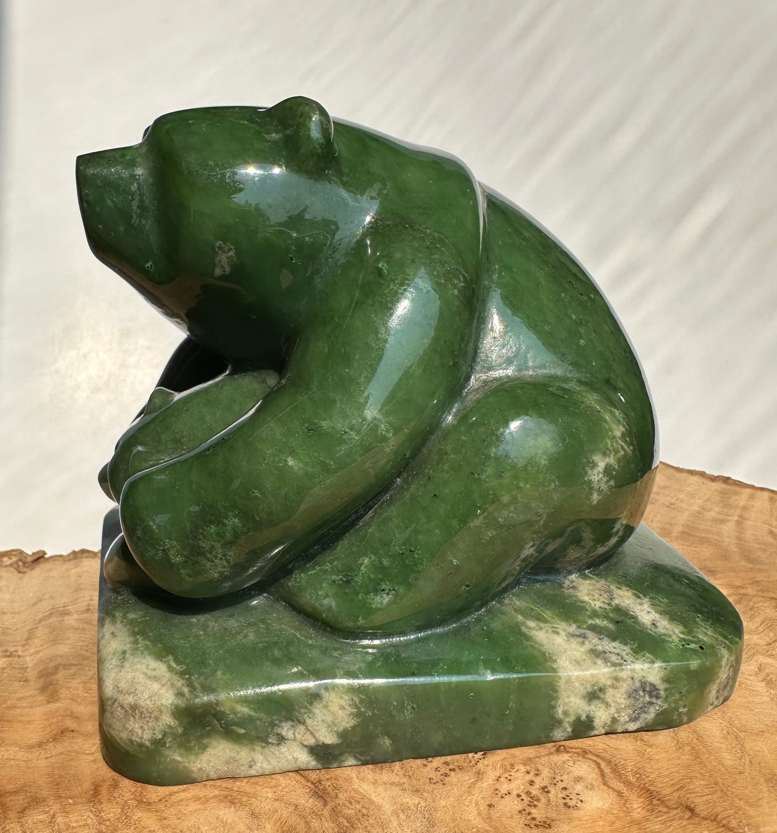 A polished green jade bear sculpture sitting on a rectangular base with natural marbled inclusions, displayed on a wood surface against a light neutral background.