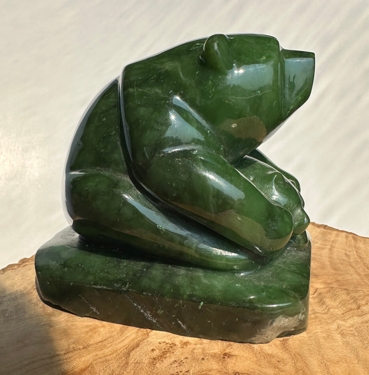 A polished dark green jade bear sculpture sitting on a natural wood slice, photographed against a neutral light background.