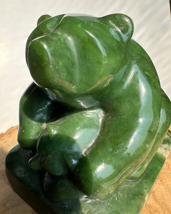A polished green jade figurine of a bear holding a cub, photographed close up against a light neutral background with a small piece of wood visible at the bottom.