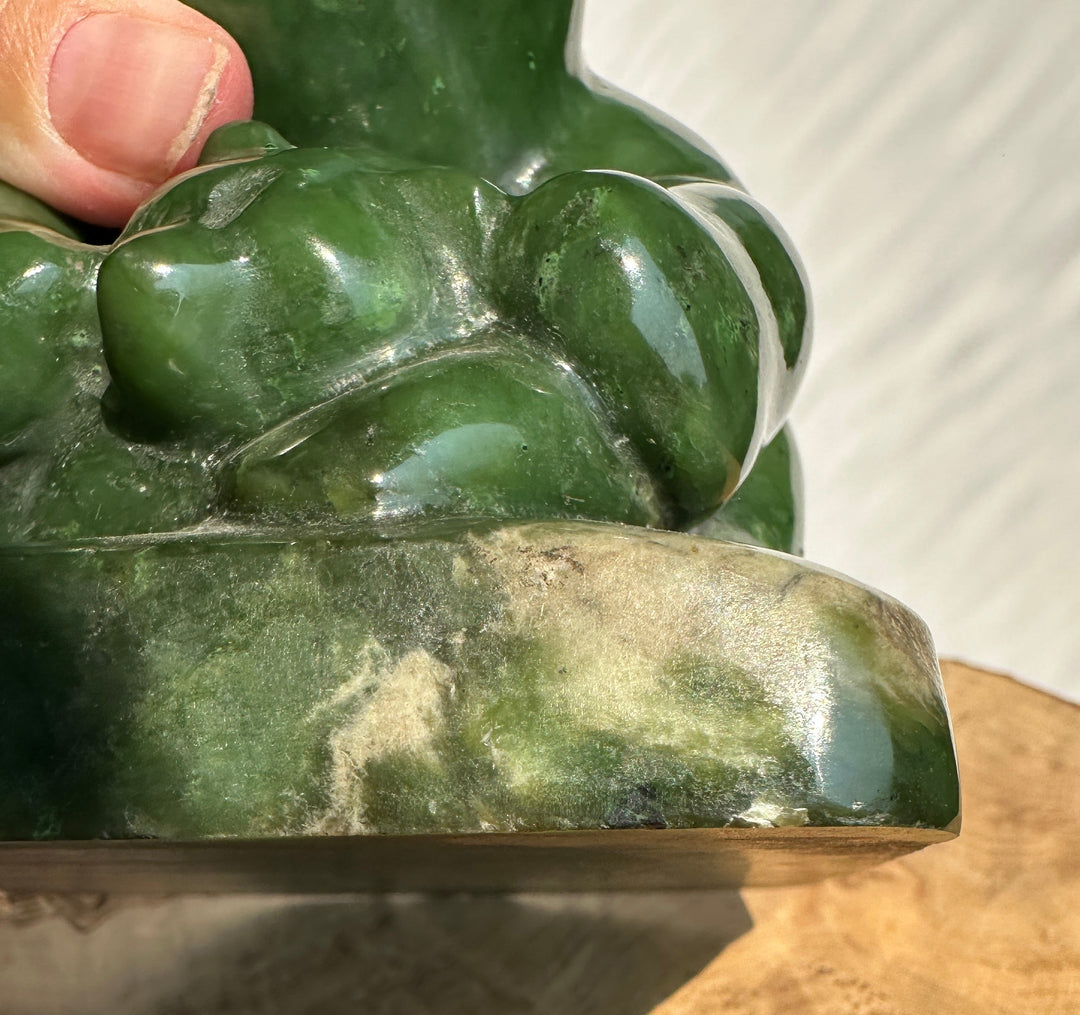 A close-up of a polished green jade figurine being held by a human hand, with the hand and a light neutral background visible.