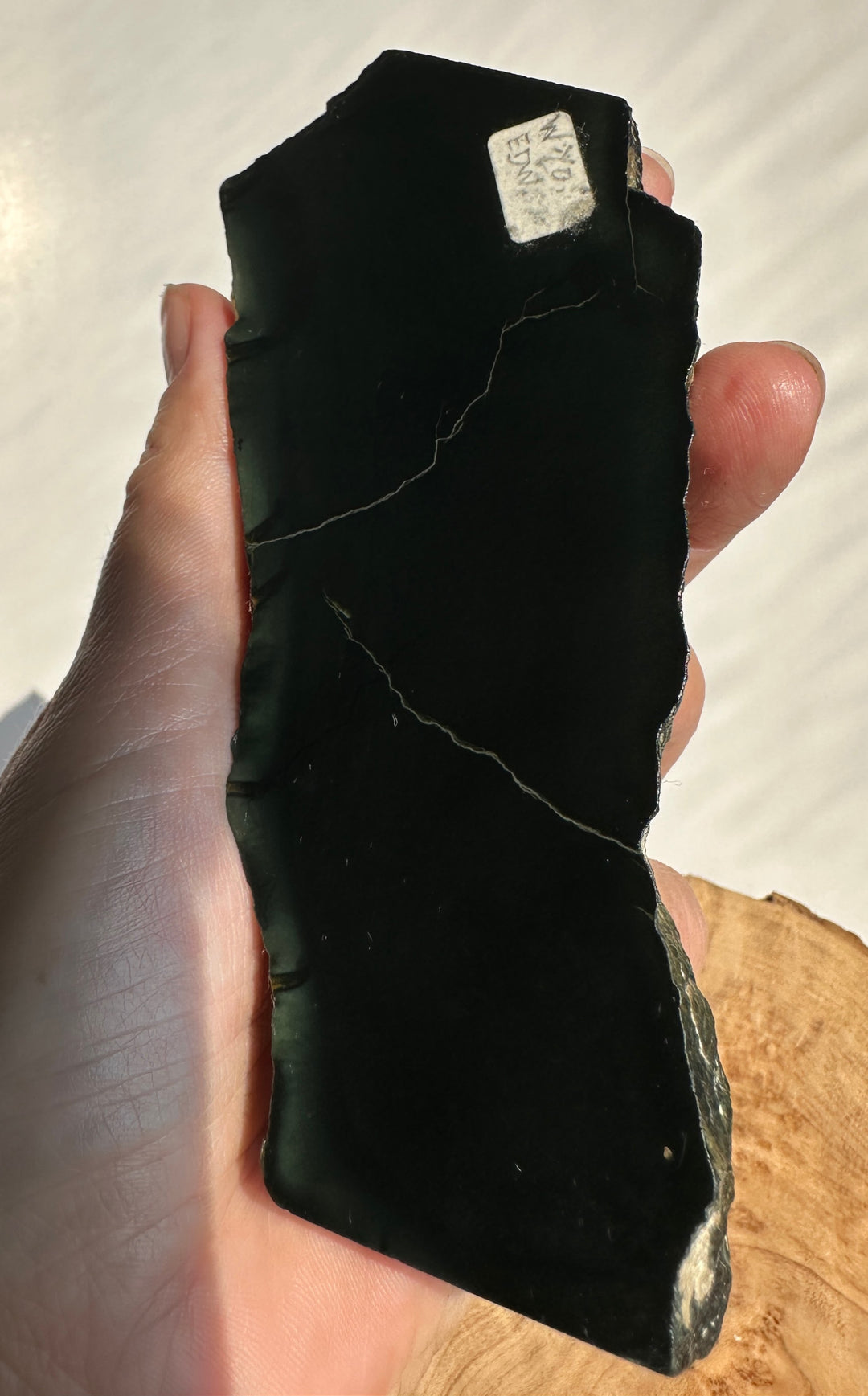 A hand holding a raw, jade  slab with natural fissures and a small white identification tag, set against a light background.