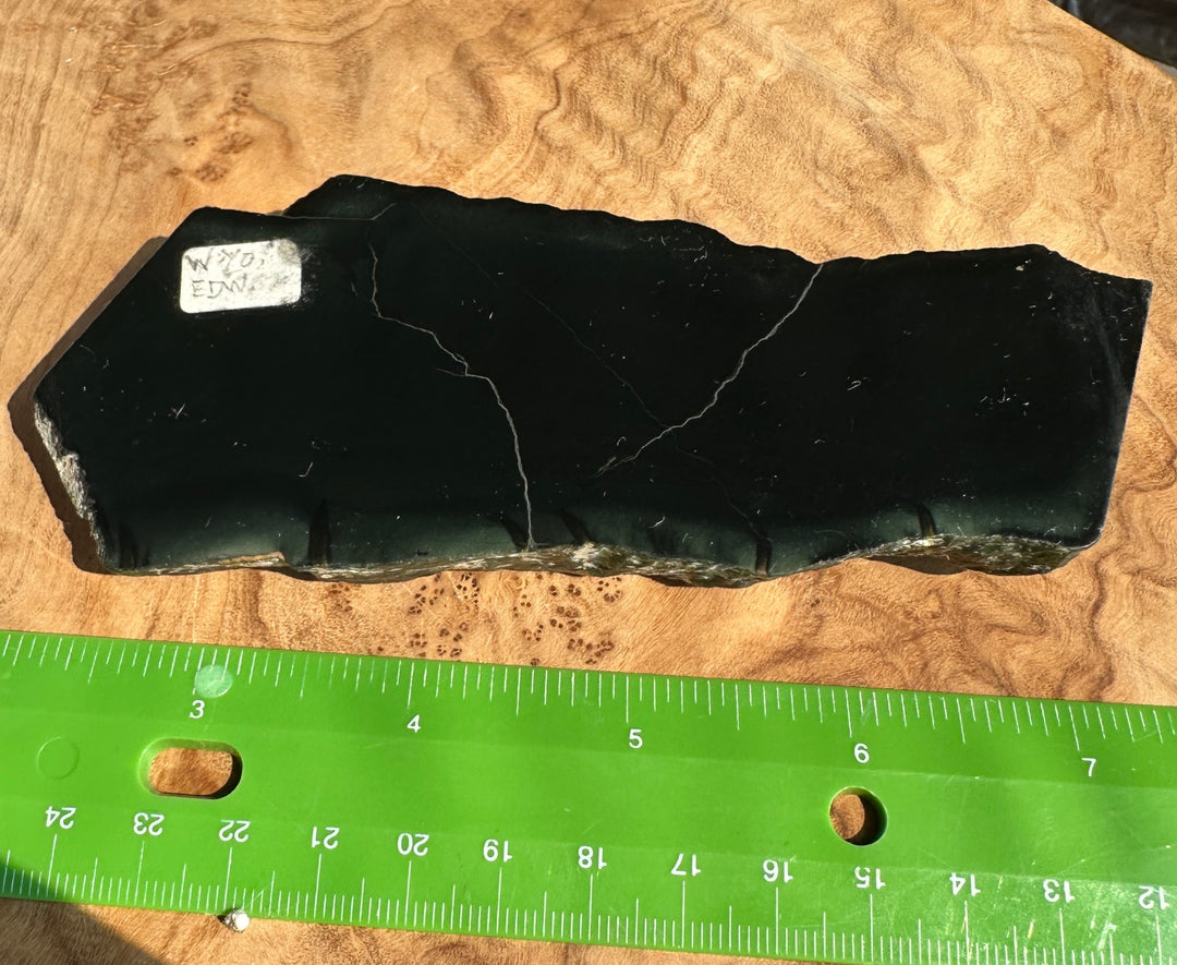 A black jade slab with natural fissures, displayed on a wood-grain surface next to a green measuring ruler for scale.