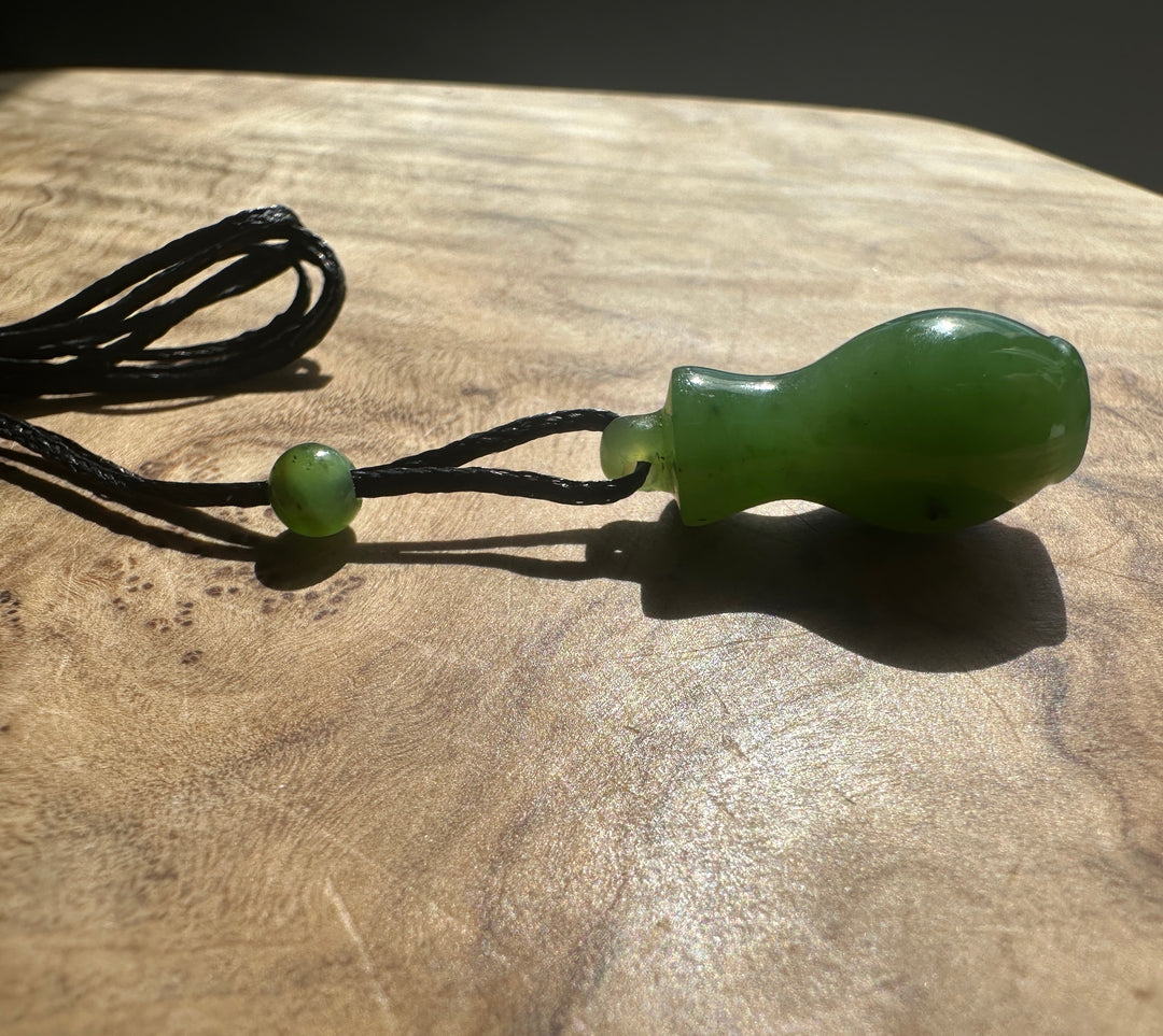 A green jade pendant with a small loop at the top, threaded on a black cord with a matching green bead, resting on a wooden surface.