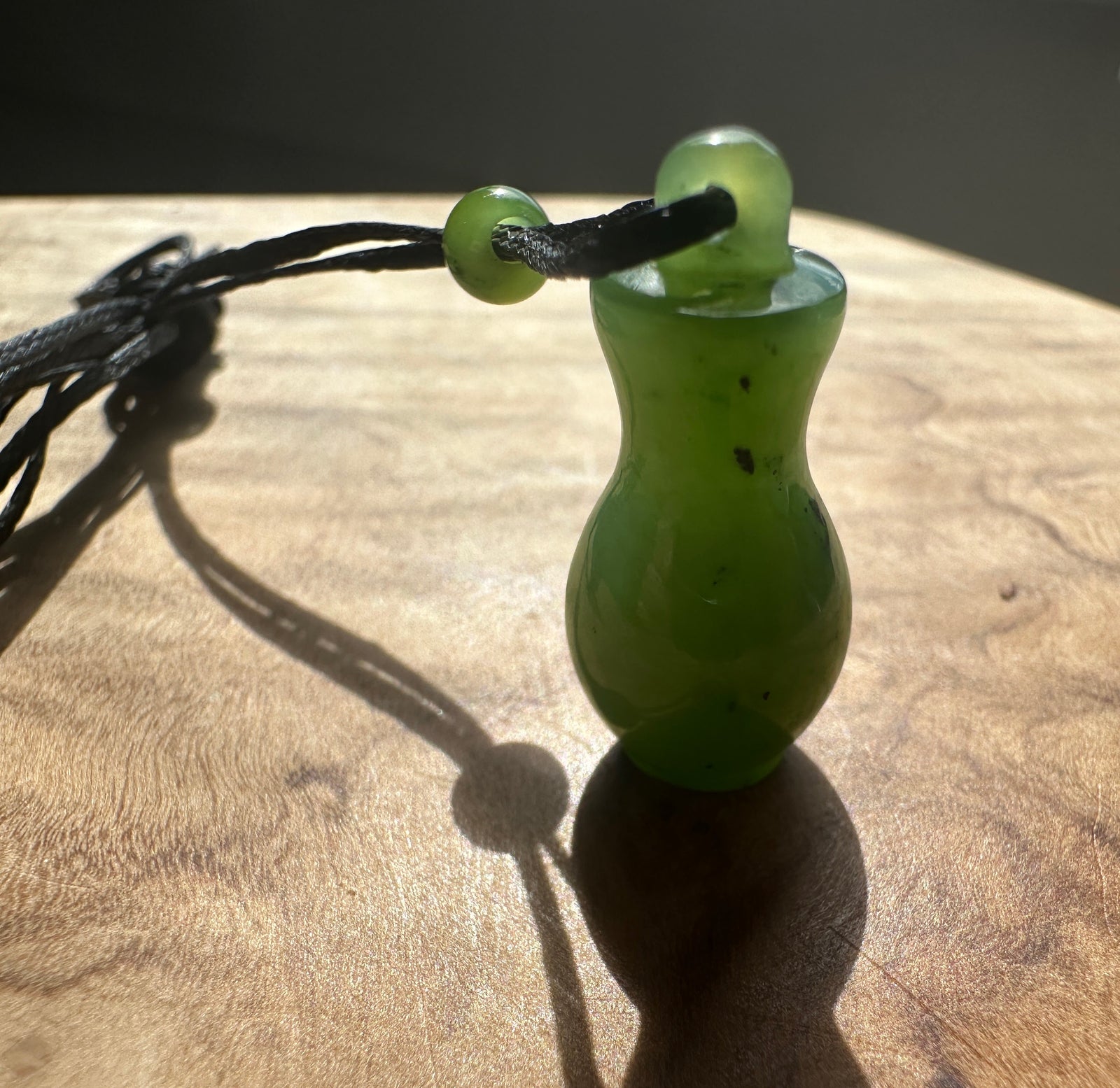 A small green jade pendant shaped like a bottle or vase, threaded on a black cord with a round green bead, resting on a wooden surface in direct sunlight.